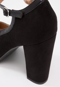 Chaussure à talon haut noire avec une finition en daim texturé et un talon large. Comprend une bride en cuir avec une boucle argentée pour la fermeture.