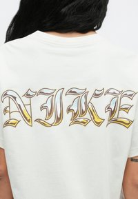 Biele bavlnené tričko s veľkým nápisom „NIKE“ v zlatej, modrej a hnedej farbe s dekoratívnym štýlom písma na zadnej strane.