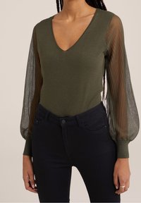 Olivgrön topp med djup V-ringning, åtsittande kropp och genomskinliga, plisserade långa ärmar; matchad med mörka skinny jeans.