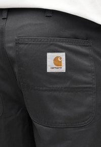 Μαύρο παντελόνι από ύφασμα με πίσω τσέπη που διαθέτει λευκή ετικέτα και πορτοκαλί λογότυπο. Το υλικό είναι απαλό με διακριτική υφή.