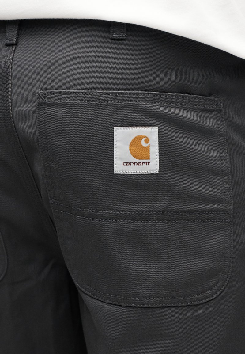 Sorte bukser i stof med en baglomme, der har et hvidt mærke og en orange logo. Materialet er glat med en diskret tekstur.