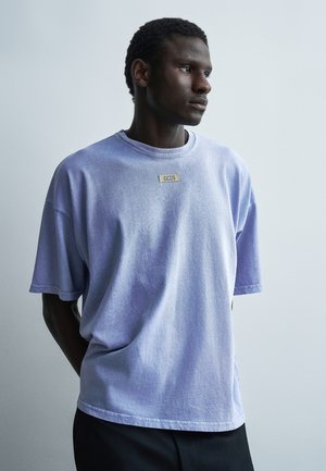 Jeune homme aux cheveux courts et bouclés portant un t-shirt ample violet clair avec une petite patch rectangulaire sur la poitrine, regardant sur le côté.