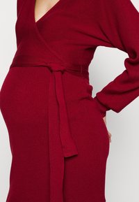 Personne enceinte portant une robe portefeuille côtelée de couleur rouge profond avec des manches longues et une ceinture nouée à la taille, la main reposant sur la hanche.