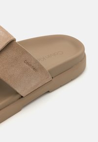 Sandale à glissière beige avec un large strap en daim. Semelle en caoutchouc et logo Calvin Klein embossé sur le strap. Texture lisse et design minimaliste.