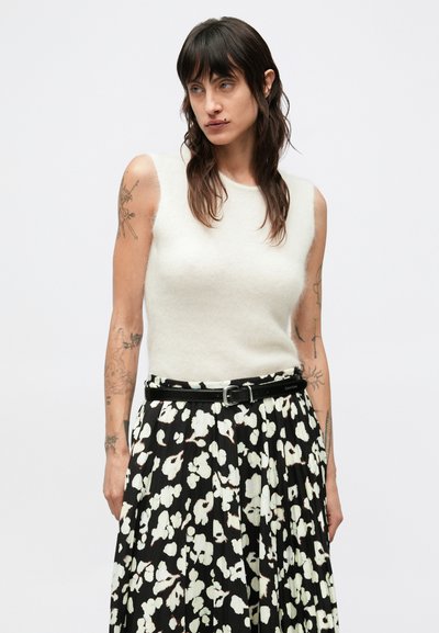 Svetlo béžový pletený top bez rukávov v kombinácii s čiernou plisovanou sukňou s kvetinovým vzorom v bielej a tlmené červenej farbe, upevnenou čiernym opaskom.