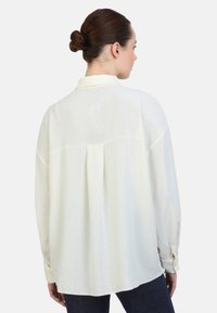 Langærmet cremebluse med en tekstureret overflade, der har en bagfold og manchetterede ærmer, parret med mørk denim.