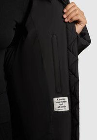 Sort quiltet jakke indvendigt med et lille hvidt label, der står "Et varmt kram lavet af stof." Har en sidelomme og en glat stofstruktur.