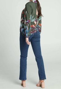 Blusa floreale verde con maniche lunghe, caratterizzata da un mix di colori vivaci. Abbinata a jeans blu scuro e sandali con tacco blocco rosa chiaro.