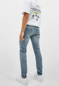 Vaqueros de mezclilla azul claro con un diseño de ajuste slim, que cuentan con bolsillos traseros y un sutil desvanecimiento, combinados con una camiseta gráfica blanca y zapatillas blancas.