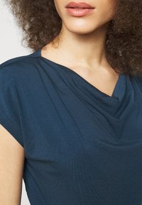 Kvinna med lockigt hår iklädd en blå topp med draperad hals och korta ärmar, som visar hennes läppar, hals och övre bröst.