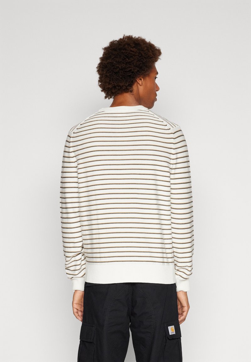 Man met krullend haar die van zich af kijkt, gekleed in een witte trui met dunne zwarte strepen en zwarte cargobroek tegen een effen achtergrond.