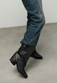 Zwarte leren enkellaarsjes met puntige neuzen en decoratieve zilveren gespen op een omgeslagen manchet. Gecombineerd met blauwe spijkerbroek.