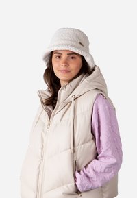 Cremfarbener Puffer-Weste mit hohem Kragen, kombiniert mit einem strukturierten, helllila Langarmshirt und einem weichen, weißen Bucket Hat.