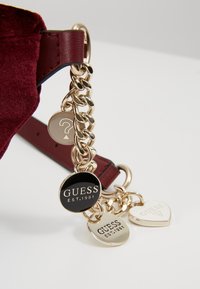 Sangle de sac bordeaux avec chaîne dorée comprenant quatre breloques : une ronde noire "GUESS EST.1981", une ronde dorée, une en forme de cœur dorée et une ronde dorée avec un point d'interrogation.