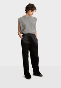 Graues ärmelloses Strickoberteil, kombiniert mit schwarzen weit geschnittenen Satin-Hosen, die über Taschen und eine glatte Textur verfügen. Die Schuhe sind schwarze Slipper.