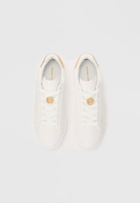 Baltas ādas sporta kurpes ar apaļu purngalu, plakaniem šņorēm, zelta logotipa akcentu uz mēles un brūnganas krāsas papēža detaļu. Gluda tekstūra, minimālistisks dizains.