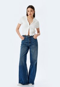Camisa blanca de textura con botones y mangas cortas, combinada con unos jeans de mezclilla azul de pierna ancha y cintura alta. Ambos artículos se exhiben sobre un fondo neutro.
