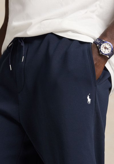 Σκούρο μπλε φόρμας από μαλακό ύφασμα, με μικρό λευκό κεντημένο λογότυπο. Περιλαμβάνει ελαστική μέση με κορδόνι και πλαϊνές τσέπες.