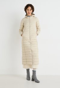 Bežasti puffer kaput s kapuljačom, dužeg kroja, kviltani dizajn, prednji zip, nosi se preko teksturirane bijele svjetne, u kombinaciji s srebrnim gležnjačama.