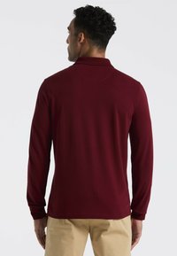 Langsleeve polo shirt in bordeaux stof met een gladde textuur. Kenmerkt een klassieke kraag en manchetten, gedragen met beige pantalon.