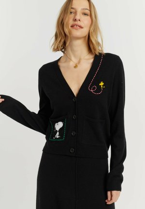 Femme portant un cardigan noir boutonné avec Snoopy sur la poche et Woodstock avec une couture rose sur la poitrine.