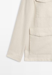 Beiger Langarmjackenärmel mit zwei vorderen Klappentaschen und sichtbaren Nähten vor weißem Hintergrund.