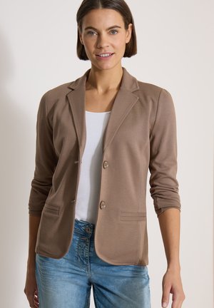 Femme portant un blazer marron clair sur un haut blanc et un jean bleu, debout devant un fond neutre avec les bras détendus.