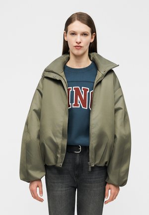 Kvinde iført oversized olivengrøn jakke over blå sweatshirt med rød tekst, mørke jeans, stående mod en ensfarvet baggrund.