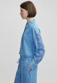 Chaqueta de mezclilla azul claro con mangas largas que presenta recortes de encaje en forma circular. Cintura con cordón y cuello tipo camisa. Textura suave.