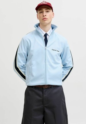 Fiatal férfi világoskék cipzáras dzsekiben fekete és fehér csíkokkal az ujján, piros sapkában, fehér ingben, sötét nyakkendővel és sötét nadrágban.