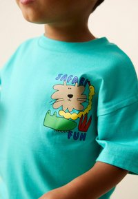 Türkisfarbenes Kurzarmhemd mit einem verspielten Tiermotiv, das einen Löwen, ein Krokodil und den Schriftzug "Safari Fun" in kräftigen Farben zeigt. Glatte Textur.