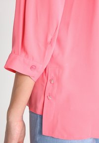 Rosa blus med korta ärmar, knapprader och en sidoklyvning. Tillverkad av ett mjukt, slätt material som passar för avslappnad användning.