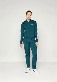 Mėlynas treningas su baltais šoniniais juostelėmis, užtrauktuku užsegama striukė ir atitinkančios kelnės. Medžiaga atrodo lygi ir sportiška, striukėje yra Adidas logotipas.