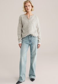 Sweater texturé gris clair avec un col rond, fermeture à boutons et coupe loose, associé à un jean taille haute bleu clair et des chaussures noires à bouts pointus.
