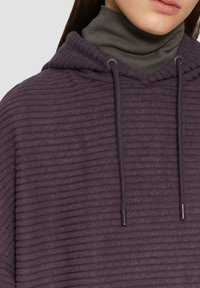Paarse hoodie met een gestructureerd, geribbeld ontwerp en een trekkoord. Gelaagd over een groene coltrui.