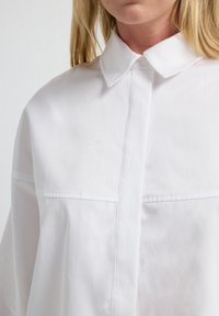 Camisa blanca de botones con cuello, que presenta una textura de tela suave, un corte relajado y costuras frontales que crean un diseño minimalista.
