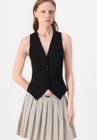 Gilet nero senza maniche con scollo a V profondo, chiusura a cinque bottoni e due tasche frontali, abbinato a una gonna plissettata beige con pois neri.