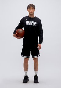 Μαύρη μπασκετική στολή που περιλαμβάνει μακρυμάνικο πουκάμισο με το κείμενο "MEMPHIS", μαύρο σορτς με λευκή επένδυση και μαύρα αθλητικά παπούτσια. Κρατάει μια μπάλα του μπάσκετ.
