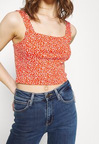 Top corto rosso con scollo quadrato, decorato con un motivo floreale bianco. Abbinato a jeans a vita alta blu scuro con chiusura a bottone.