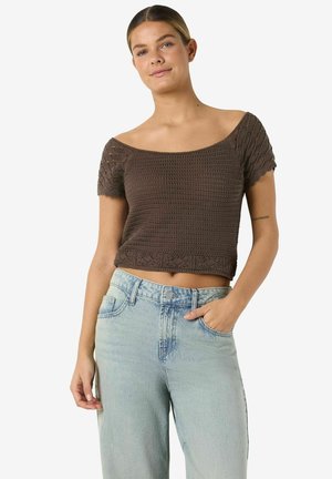 Ung kvinne iført en brun off-shoulder hekle-topp og lyseblå høytlivsjeans, står med en hånd i lommen.
