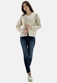 Crème ribbel cardigan met een V-hals en striksluitingen, gedragen over een witte T-shirt, gecombineerd met donkerblauwe jeans en witte sneakers.