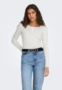 Hvit ribbestrikket langermet topp med rund hals og en knepping med tre knapper. Kombinert med lyseblå høytlivs jeans og en svart belte.