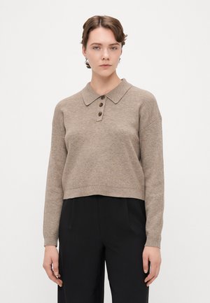 Pull polo tricoté marron avec col, trois boutons marron et ourlet court. Porté avec un pantalon noir plissé.