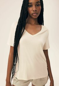Beżowy t-shirt z dekoltem w serek, wykonany z miękkiego, lekkiego materiału, z krótkimi rękawami. Nosi go z wzorzystymi szarymi szortami z małymi białymi kropkami.