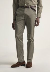 Hombre vestido con pantalones verde oliva, una camisa de manga larga a juego, un cinturón trenzado marrón y mocasines de ante marrón sobre un fondo claro.