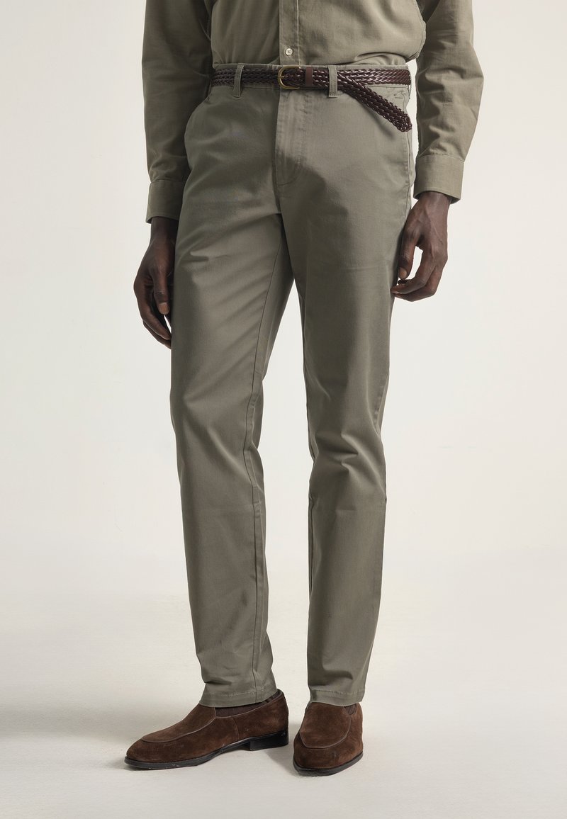 Hombre vestido con pantalones verde oliva, una camisa de manga larga a juego, un cinturón trenzado marrón y mocasines de ante marrón sobre un fondo claro.