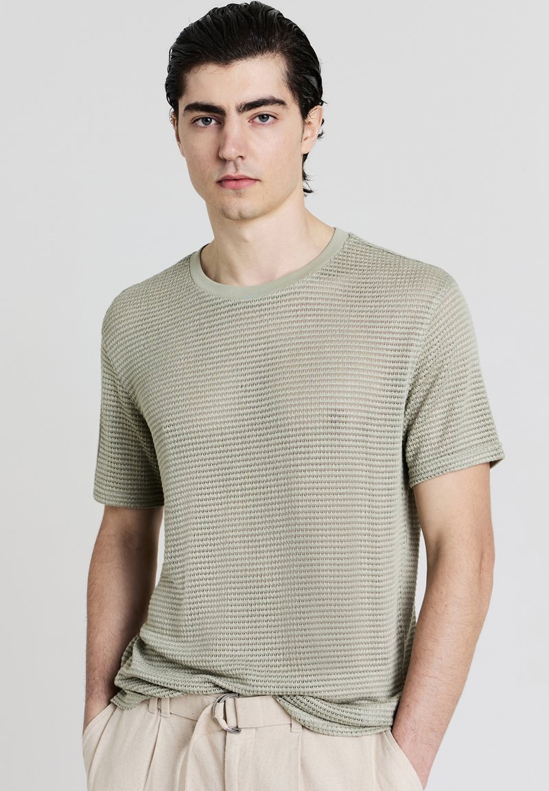 Giovane uomo con capelli scuri, indossa una camicia a maniche corte di colore verde chiaro con texture e pantaloni beige, in piedi davanti a uno sfondo chiaro e semplice.