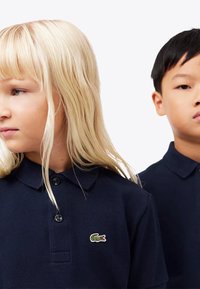 Kaksi lasta, joilla on tummansiniset polo-paidat, joissa on pieni vihreä krokotiililogo, kääntyneenä vastakkaisiin suuntiin valkoista taustaa vasten.