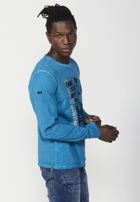 Long-sleeve blue shirt with a textured fabric, featuring a black graphic design on the front. Loose fit, round neckline, and visible stitching.