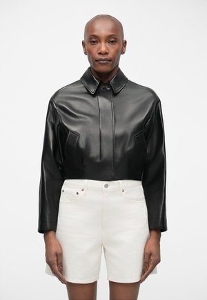 Veste en cuir noire cropped avec un col, présentant des détails de couture à l'avant, associée à des shorts en denim blancs. Design épuré et texture lisse.